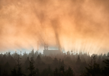Sun rises, its rays pierce through the mist, casting dramatic beams of light over landscape. Antenna tower and building emerge from fog, surrounded by dense forest of pine trees.の写真素材