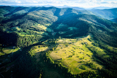 Aerial view of sunlit valley, dotted with small houses and winding roads, surrounded by dense forests. Golden light casts long shadows. Vibrant landscape stretches out, merging into forested hills.の写真素材