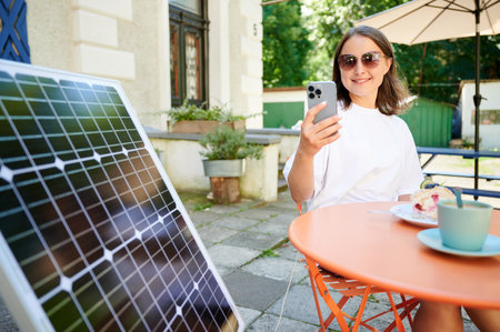 Young woman charging her smartphone from solar panel while relaxing. Pretty girl drinking coffee and checking her phone next to solar battery. Conscious female choosing sustainable lifestyle.の写真素材