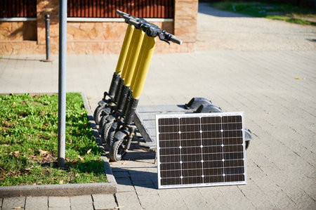 Row of five electric scooters with yellow handlebars parked on sidewalk next to patch of grass. Solar panel placed nearby, indicating eco-friendly sustainable charging solution for scooters.の写真素材