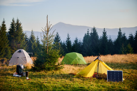 Solar panel in grassy field with tents in background highlights eco-friendly camping setup, surrounded by dense evergreen trees in evening. Integration of renewable energy in outdoor activities.の写真素材
