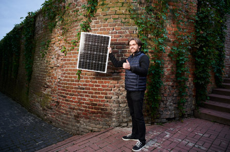 Man holding photovoltaic solar panel in front of old brick wall covered in green ivy, giving thumbs up. Concept of integration of sustainable renewable energy sources into historical architecture.の写真素材