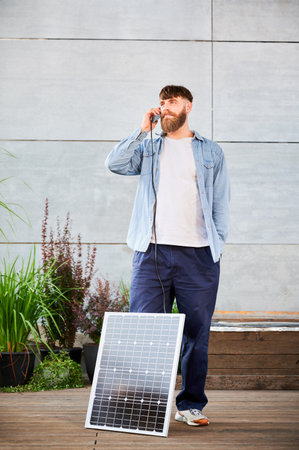 Bearded man talks on smartphone connected to photovoltaic solar panel. Integration of sustainable renewable energy into everyday life, demonstrating practical use of solar power for charging devices.の写真素材