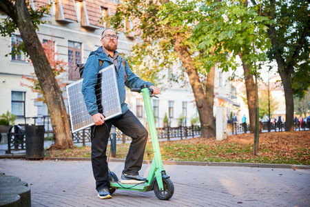 Man rides electric scooter while carrying solar panel. Integration of solar power as sustainable energy source for charging electric scooters, promoting eco-friendly urban transportation.の写真素材