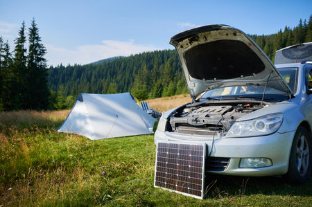 Solar panel in front of car with hood open, symbolizing transition from traditional internal combustion engines to electric cars. Charging cable connects solar panel to vehicle, renewable energy usageの写真素材