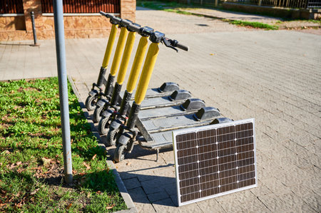 Row of five electric scooters with yellow handlebars parked on sidewalk next to patch of grass. Solar panel placed nearby, indicating eco-friendly sustainable charging solution for scooters.の写真素材
