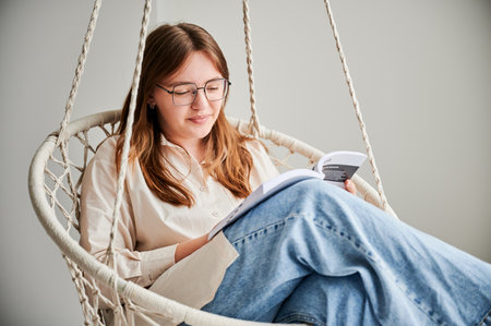 Beautiful girl relaxing and reading book on indoor swing. Pretty female in glasses excited about story she reading. Cozy afternoon with book in hanging chair in home.の写真素材