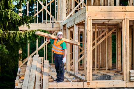 Carpenter constructing wooden-framed house near the woods. Bearded man with spectacles holding large joist, wearing work clothes and helmet. Concept of modern and eco-friendly building.の写真素材