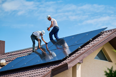 Workers building solar panel system on rooftop of house. Two men installers in helmets installing photovoltaic solar module outdoors. Alternative, green and renewable energy generation concept.の写真素材