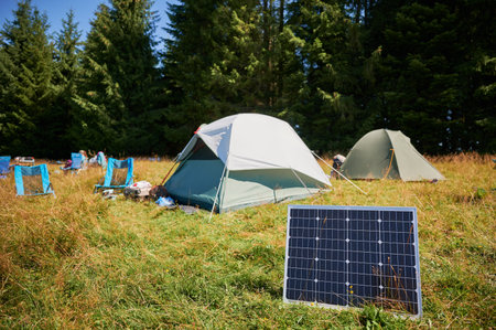 Solar panel in grassy field with tents in background highlights eco-friendly camping setup,...