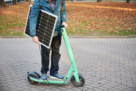 Cropped view of man rides electric scooter while carrying solar panel. Integration of solar power as sustainable energy source for charging electric scooters, promoting eco-friendly transportation.の写真素材