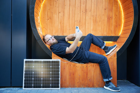 Bearded man using smartphone connected to photovoltaic solar panel. Integration of sustainable renewable energy into everyday life, demonstrating practical use of solar power for charging devices.の写真素材