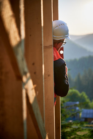 Carpenter constructing wooden frame house. Back view of man, wearing spectacles, inspects the walls for levelness using spirit level. Concept of environmentally-friendly contemporary construction.の写真素材