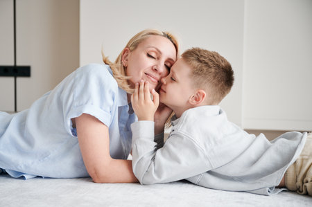 Little son showing love to his mom. Young mother and her kid lying on white carpet and spending time together at home. Lovely afternoon with family.の写真素材