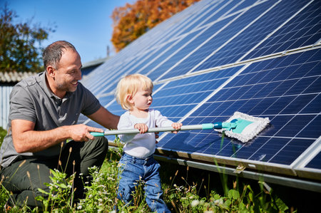 Father showing his little son the solar panels during sunny day. Cute boy helping his dad to clean solar battery. Young man teaching his small kid how to care about their source of energy.の写真素材