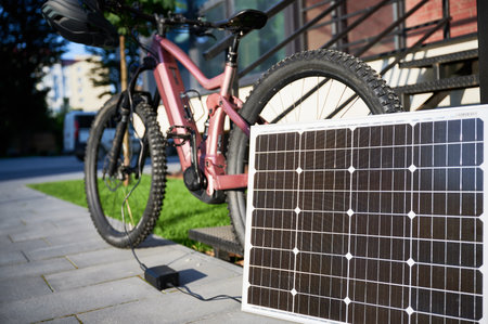 Pink electric mountain bike parked beside staircase, connected to small solar panel for charging. Bike rugged tires, mounted helmet on handlebar, situated on paved pathway with grassy lawn.の写真素材