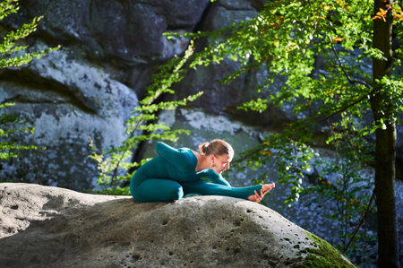 Woman practicing yoga outdoors in forest. Barefoot female on yoga mat surrounded by trees and large rocks, which suggests peaceful, natural environment ideal for meditation or yoga practice.の写真素材