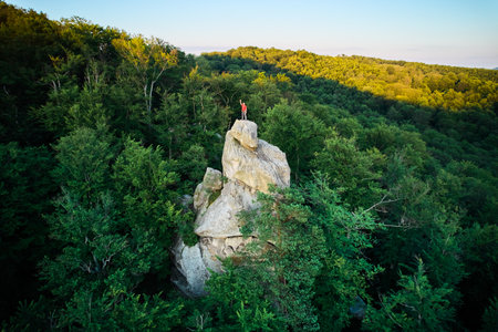 Aerial view of climber standing atop rock formation at Dovbush Rocks in Carpathian mountains, Ukraine. Sun sets, casting warm glow over lush, green forest, distant hills, creating breathtaking view.の写真素材