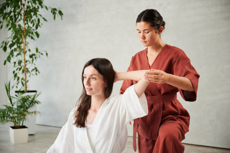 Masseuse making thai yoga massage. Woman therapist doing traditional massage treatment for female patient indoors. Client sitting, meditating, enjoying hand massage in calm atmosphere.の写真素材