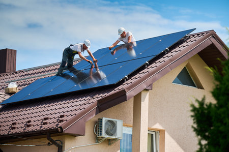 Workers building photovoltaic solar panel system on rooftop of house. Men technicians in helmets installing solar module with help of hex key outdoors. Renewable energy generation concept.の写真素材
