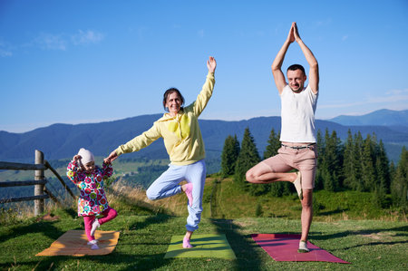 Young family doing yoga outdoors in scenic mountainous landscape in morning. Father, mother and daughter on yoga mats, with scenic view of mountains and forest in background under clear blue sky.の写真素材
