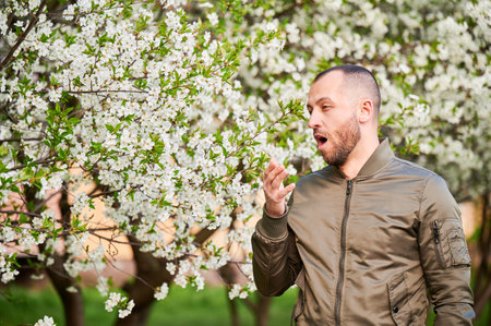Man allergic suffering from seasonal allergy at spring in blossoming garden at springtime. Bearded young man sneezing and having runny nose in front of blooming tree. Spring allergy concept.の写真素材