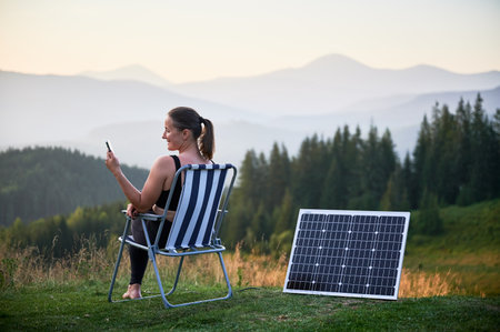 Back view of young happy woman sits in chair, using smartphone beside solar panel. Landscape of rolling hills and mountains. integration of technology with nature for eco-friendly experience.の写真素材