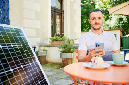 Young man charging smartphone from solar panel while sitting in cafe. Handsome male drinking coffee and checking his phone next to solar battery. Conscious male choosing sustainable lifestyle.の写真素材
