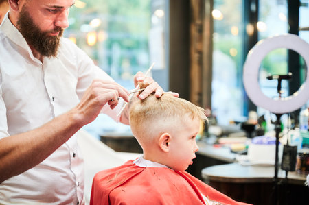 Man barber cutting little boys hair using comb and scissors. Child getting haircut from adult male, likely barber. Professional hairdresser and cute client at modern barbershop.の写真素材