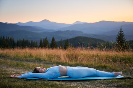 Woman practicing yoga outdoors in the mountains in a serene, natural setting. Female performing yoga pose on blue mat, with backdrop of beautiful mountain landscape at sunrise or sunset.の写真素材