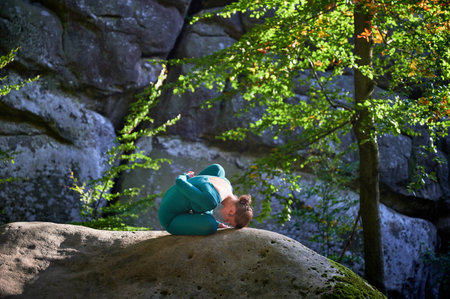 Woman practicing yoga outdoors in forest. Barefoot female on yoga mat surrounded by trees and large rocks, which suggests peaceful, natural environment ideal for meditation or yoga practice.の写真素材
