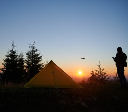 Peaceful campsite at sunrise with vibrant yellow tent pitched among pine trees. Silhouette of person operating drone adds dynamic element to serene scene, as sun casts golden glow across horizon.の写真素材