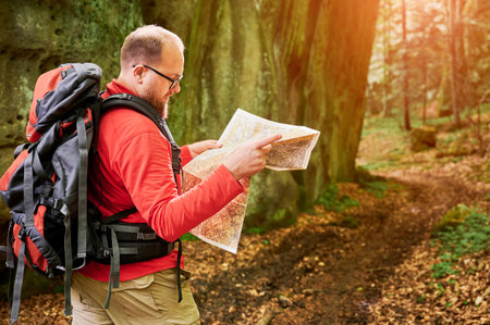 Bearded tourist man studies map while stands in front of large rock in dense forest. Traveler with grey backpack and glasses, planning outdoor adventure, hiking or climbing route.の写真素材