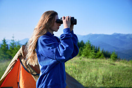 Woman stands near orange tent on grassy hilltop, looking through binoculars, wearing blue hoodie. Female hiker observes distant mountains and lush green forest under clear blue sky.の写真素材