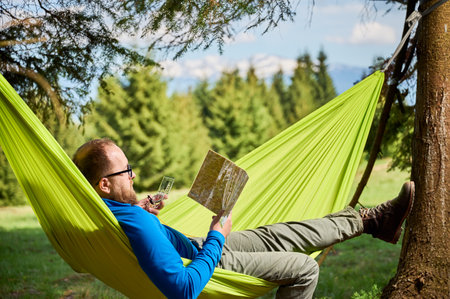 Man traveler in glasses studies map while holding compass, planning outdoor adventure or hiking route, relaxes in green hammock in forested area. Trees with distant mountains on blurred background.の写真素材