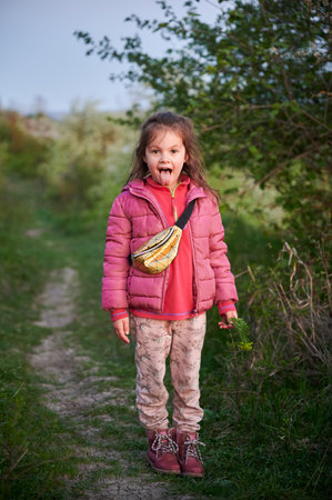 Young girl playfully sticks out her tongue while standing on a nature trail. Child dressed in pink jacket, red shirt, patterned leggings, and red boots, with small crossbody bag.の写真素材