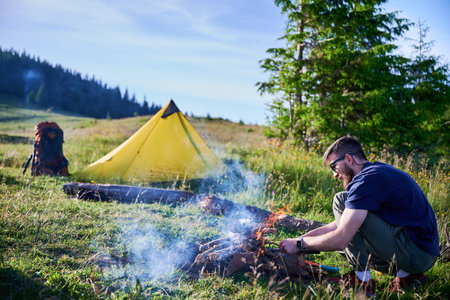 Man tending to campfire on grassy field with yellow tent and backpack nearby. Smoke rises from fire as he works, surrounded by lush greenery and distant tree line under clear blue sky.の写真素材