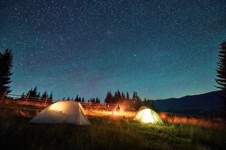 Silhouette of couple hikers resting by campfire under star-studded sky, near illuminated tents at campsite. Night camping in mountains, night sky create magical and peaceful atmosphere.の写真素材
