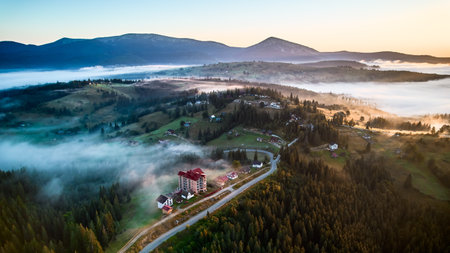 Aerial view of misty morning in mountains, with small village nestled among trees. Fog gently blankets landscape, creating serene and picturesque scene, as sun begins to rise in the background.の写真素材