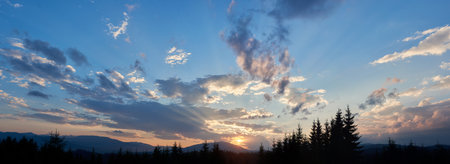 Sun sets behind range of distant mountains, casting golden rays through clouds. Sky, painted with shades of blue, pink, and gold, in tranquil evening. Silhouetted pine trees add depth to landscape.の写真素材