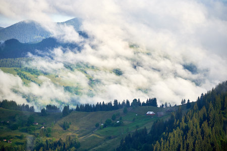 Picturesque scene of mountain valley blanketed in mist, with patches of green pastures and scattered houses. Clouds drift gently over forested hills, creating serene and tranquil atmosphere.の写真素材