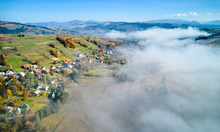 Picturesque village nestled in rolling hills, bathed in autumn colors with patches of green and yellow fields. Wisps of fog drift through landscape, adding dreamy atmosphere to serene rural scene.の写真素材