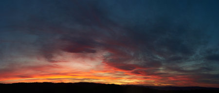 Stunning sunset paints sky with vibrant hues of red, orange, and pink, blending into deep blues and purples. Silhouetted hills create contrast against fiery horizon, adding drama to serene landscape.の写真素材
