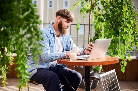 Man works on laptop and smartphone at outdoor table, with glass of iced coffee. Solar panel nearby, emphasizing sustainable, modern eco-friendly workspace that combines technology and sustainability.の写真素材