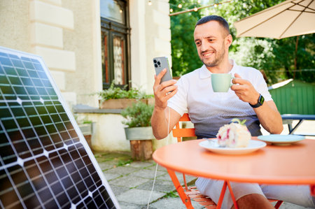 Young man charging smartphone from solar panel while sitting in cafe. Handsome male drinking coffee and checking his phone next to solar battery. Conscious male choosing sustainable lifestyle.の写真素材