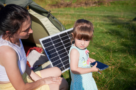 Young child and mother play with smartphone while charging with photovoltaic solar panel near tourist tent in summer. Integration of renewable energy in outdoor camping activities.の写真素材