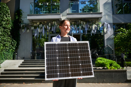 Woman holding solar panel for charging, stands outdoors, smiling in urban setting. Concept of green energy generation.の写真素材