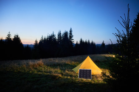 Yellow tent with solar panel in grassy field at twilight, surrounded by dark silhouettes of trees. Solar panel emphasizes eco-friendly setup, capturing tranquility of nature as night falls.の写真素材