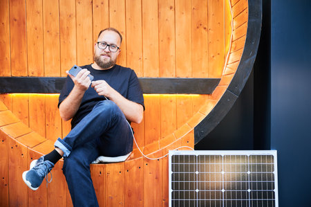 Bearded man connecting smartphone to photovoltaic solar panel. Integration of sustainable renewable energy into everyday life, demonstrating practical use of solar power for charging devices.の写真素材