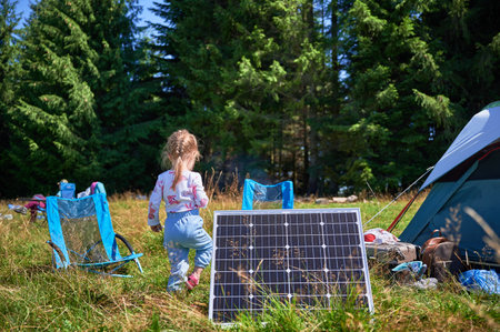 Young child playing near tourist tent with photovoltaic solar panel in summer. On background of dense forest. Integration of renewable energy in outdoor camping activities.の写真素材